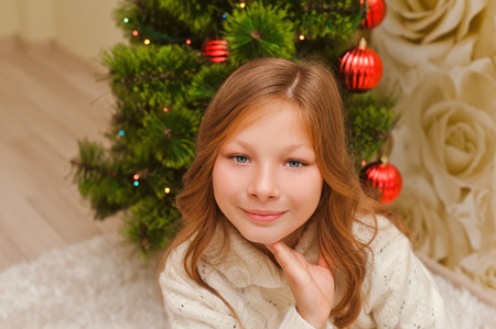 Girl sitting on a light carpet, smiling, looking at the camera. Child with long hair. Christmas tree. Teenager in a warm white sweater.の写真素材