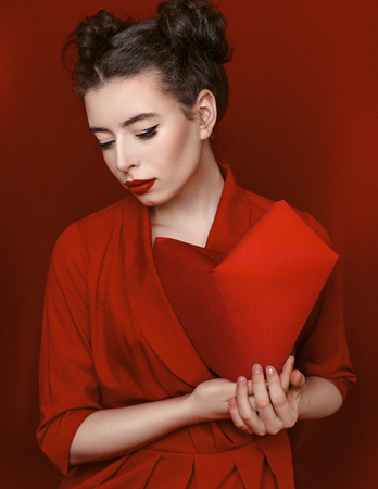 Brunette young woman with red lips wearing red dress on red background. Girl is posing in studio for Valentines day greeting card.の写真素材