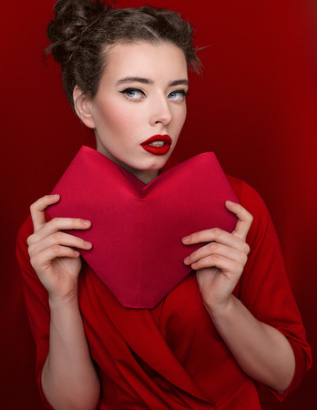 Brunette young woman with red lips wearing red dress on red background. Girl is posing in studio for Valentines day greeting card.の写真素材