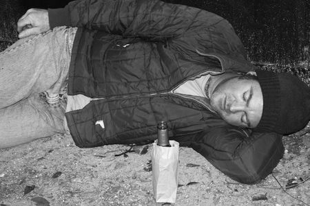 A black and white photo of a homeless man sleeping on the ground by a dumpster.  He has a bottle of wine in a bag beside him.の写真素材