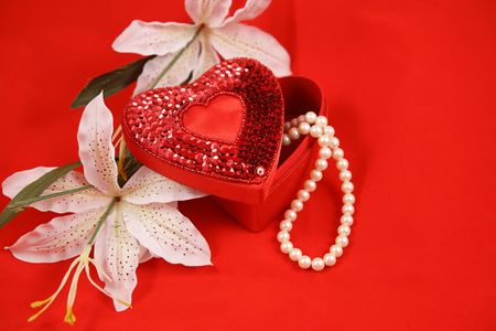 A heart shaped box with a pearl necklace, and easter lilies on a red background. Room for text.の写真素材
