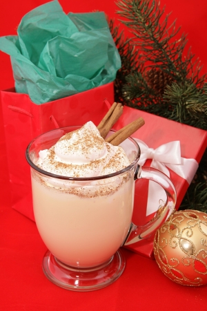 A frothy mug of Christmas eggnog with a dollop of whipped cream, nutmeg and cinnamon sticks, nestled among Christmas gifts and decorations.  Red background.の写真素材