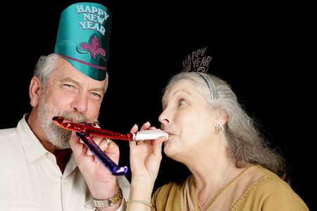 Mature couple in New Year's party hats blowing noise-makers.  Isolated on black.  の写真素材
