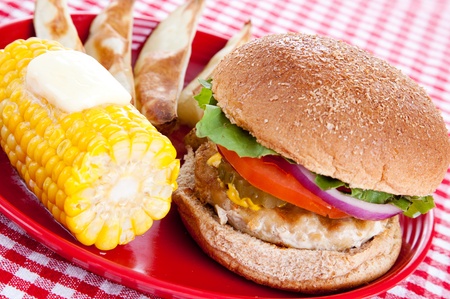 Healthy turkey burger on whole grain bun, with baked potato wedges and corn on the cob, against a red and white checked table cloth.  の写真素材