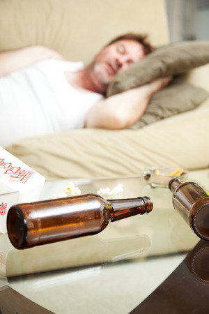 Man asleep on the couch with ashtray, takeout food container, and beer bottles on coffee table.  Shallow Depth of field with focus on beer bottles.  の写真素材