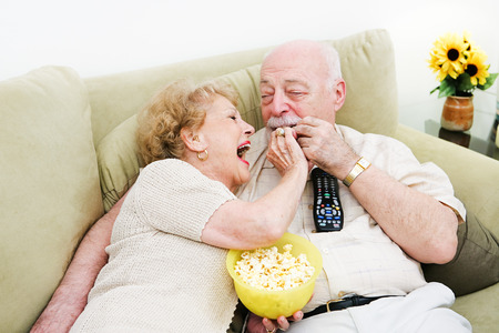 Senior woman laughs and feeds popcorn to her husband as they watch television.の写真素材