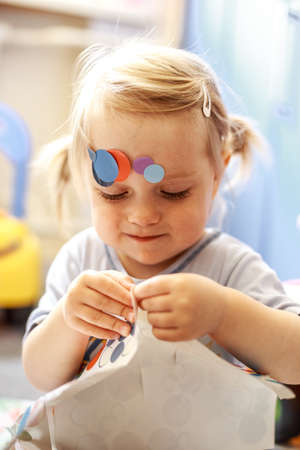 Portrait of a little lovely girl. Cute toddler sticks stickers on herself. Lovely blonde baby in a blue shirt.の写真素材