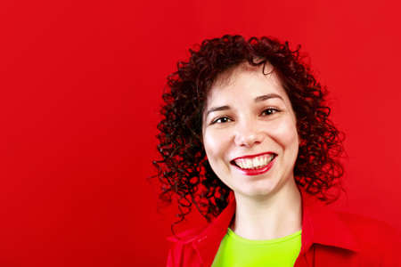 Studio shot of positive and the smiling female. Curly girl in bright shirt and salad t-shirt. Isolated on red background.の写真素材