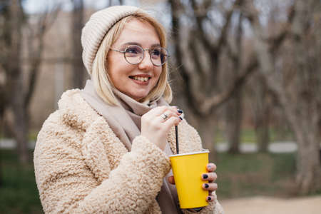 Pretty casual blonde in the park, smiling and drinking coffee to go. Young woman in modern wear...