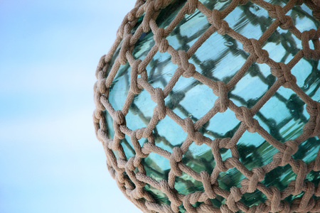 Turquoise green sea glass sphere tied up with nautical rope & knots against a blue sky.