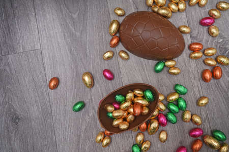 Pile or group of multi colored and different sizes of colourful foil wrapped chocolate easter eggs in pink, red, gold and green. Large halves of a brown milk chocolate egg have mini eggs inside, on a grey wooden background.の写真素材