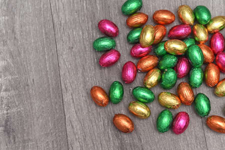 Foil wrapped multi coloured easter eggs in pink, green, orange and yelow in a pile or group, against a grey white wooden background.の写真素材