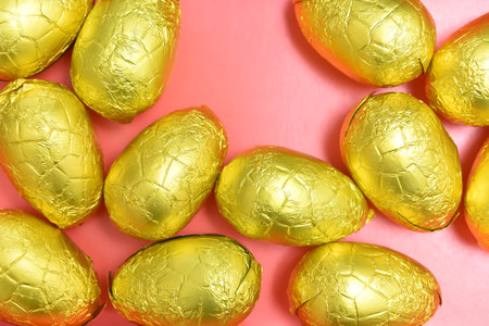 Pile or group of gold, or yellow foil wrapped chocolate easter eggs, against a pink, peach orange...