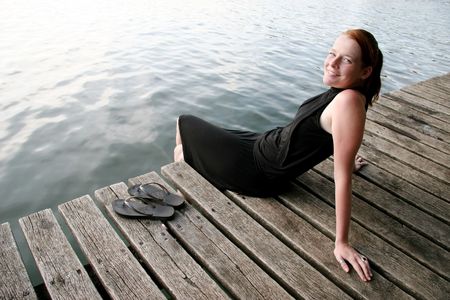 A young woman in a black dress relaxing on a wooden peer at a lake.の写真素材