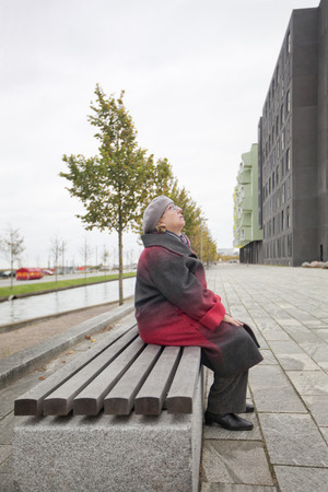 An elderly woman in a red coat is resting on a bench, on the street and looking upの写真素材