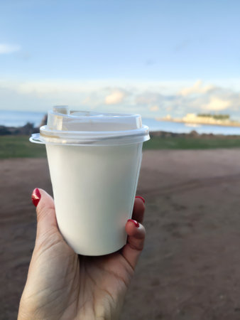 A womans hand holds a paper cup of coffee against the backdrop of the Gulf of Finland. Close-up of a...