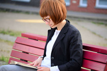 young woman with glasses sits on a bench and reads a book