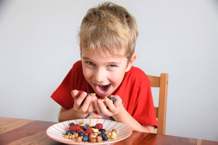 Joyful boy holds berries and nuts in his palms over a plateの写真素材