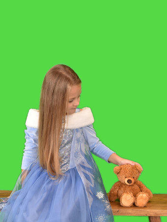 a girl with long hair in a beautiful blue dress sits on a wooden bench and strokes a teddy bearの写真素材