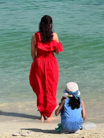 a woman in a red dress stands on the seashore and a girl sits next to herの写真素材