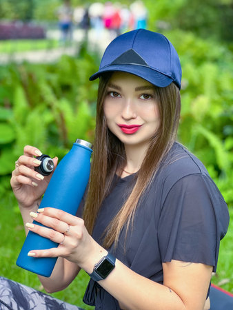 Young woman with long hair in a cap holding a metal beverage bottle outdoorsの写真素材