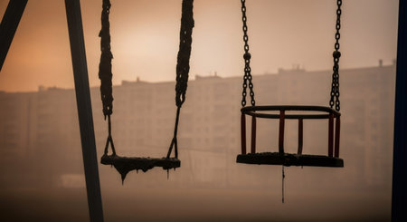 Empty dirty swings on a playground with distant buildings in thick brown haze. Concept of environmental pollution, disaster, and abandonment.の素材