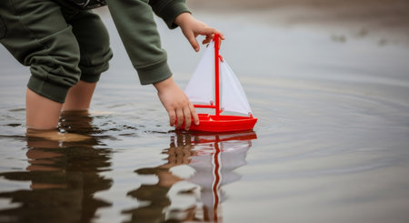 Child plays with red toy sailboat in water. Barefoot kid having fun launching small ship in shallow puddle. Outdoor active game.の素材