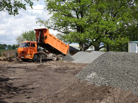 Construction equipment an orange dump truck unloads crushed stone and sand at the site of a village...