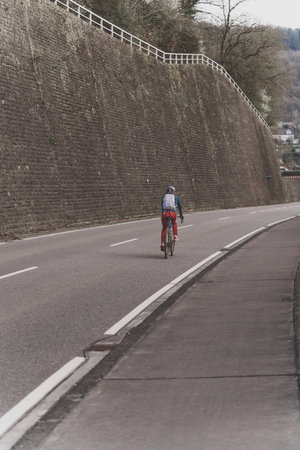A solitary cyclist traverses a winding road, accompanied by a towering stone wall and lush greenery aroundの写真素材