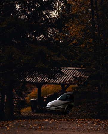Car parked under wooden shelter dusk, moody autumn forest scene with gravel driveway and scattered...