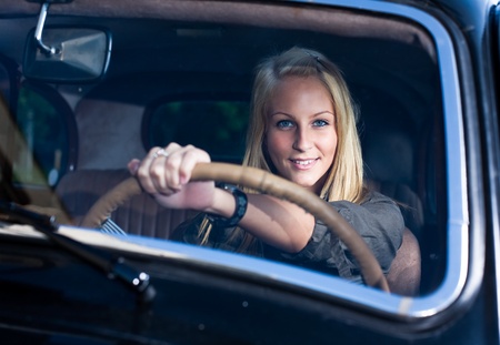 Beautiful young blond girl smiling in a black vintage car, holding the streering wheel, shot through the windscreen.の写真素材