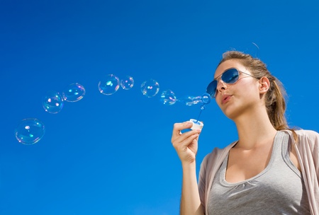Beautiful young brunette girl blowing a wave of soap bubble to the cear blue sky.