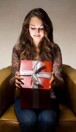Portrait of a beautiful young brunette peeking inside shiny red gift box in creative lighting.の写真素材
