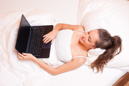 Beautiful young brunette woman with her laptop in bed.の写真素材