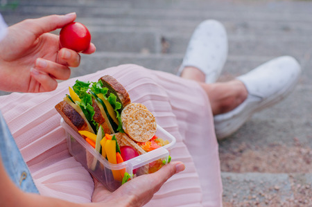 Healthy food concept: Young woman eating from lunch box filled with sandwich, crispbreads, fruits and vegetables sitting on stairs; selective focus on lunchboxの写真素材