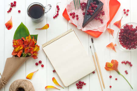 Open empty notebook with cup of coffee, red current cake slice, brushes and bouquet of flowers in the craft paper on the white wooden background, copy space. Top view.の写真素材