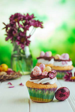Happy Easter card: Easter vanilla cupcakes decorated with candy eggs in nest on white wooden...
