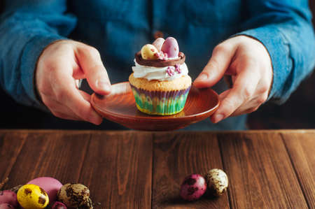 Man's hands holding Easter vanilla cupcake decorated with candy eggs in nest over rustic wooden...