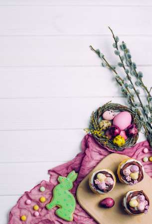 Happy Easter card: Easter nest cupcakes, Easter rabbit gingerbread, painted eggs in natural nest and blooming pussy-willow brunches on white wooden background with copy space top view, flat layの写真素材