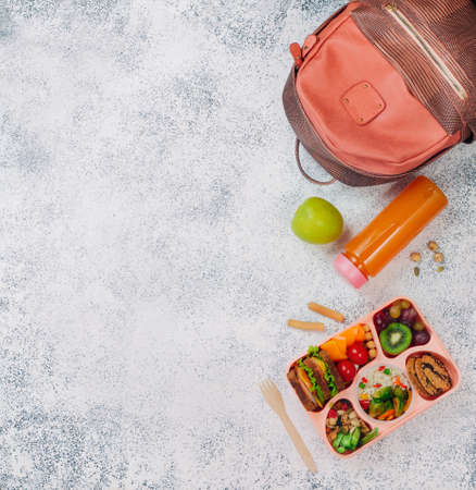 Healthy food concept: Open lunch box with sandwiches, vegetable salad, fresh grape and kiwi, rice with grilled Brussels sprouts and nuts on the gray background near backpack with copy space top view, flat layの写真素材