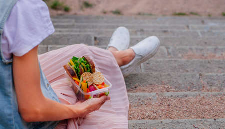 Young hipster girl eating from lunch box filled with sandwich, crispbreads, fruits and vegetables...