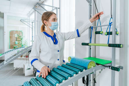Female doctor rehabilitologist wearing protective face mask showing training training apparatus