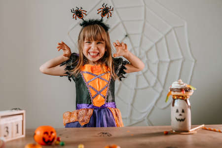 Portrait of little girl dressed Halloween witch costume and spider headband sitting at the table with sweets and monster shake or freak milkshake. Selective focus.の写真素材