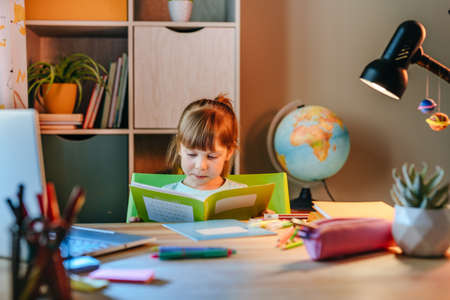 Elementary female student studying online from home using laptop sitting at the desk in the room. Distance education during pandemic lockdown concept.の写真素材