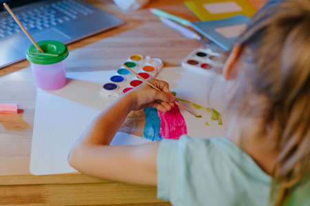 Elementary female student studying online drawing from home using laptop sitting at the desk in the room. Distance education during pandemic lockdown concept.の写真素材