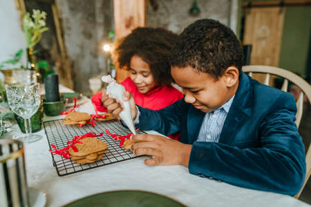 Sister and brother decorating gingerbreads with ribbons sitting at the table at home. Christmas time family leisure concept. Idea for diy handmade Happy New Year sweets, gift or tree decoration.の写真素材