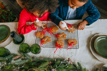 Sister and brother decorating gingerbreads with ribbons sitting at the table at home. Christmas time family leisure concept. Idea for diy handmade Happy New Year sweets, gift or tree decoration.の写真素材