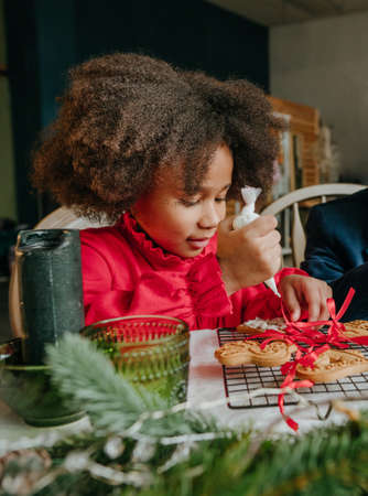 Girl decorating gingerbreads with ribbons sitting at the table at home. Christmas time kids leisure concept. Idea for diy handmade Happy New Year sweets, gift or tree decoration.の写真素材