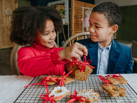 Sister and brother presenting each other gingerbreads with ribbons sitting at the table at home. Christmas time concept. Idea for diy handmade Happy New Year sweets, gift or tree decoration.の写真素材