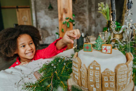 Girl taking gingerbread from Christmas cake decorated with handmade house gingerbreads. Idea for diy handmade Happy New Year festive sweets. Selective focus on the cake.の写真素材